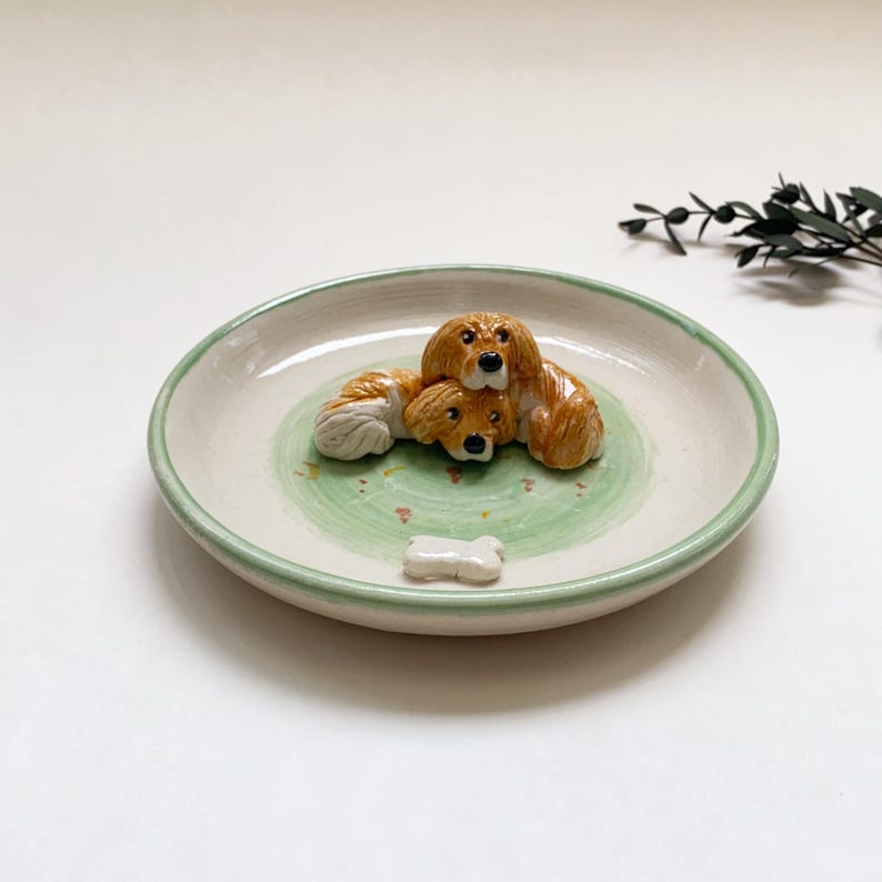 May include: A ceramic decorative plate with a light green rim and a green and white interior. The plate features three brown and white dog figurines and a small bone-shaped figurine.