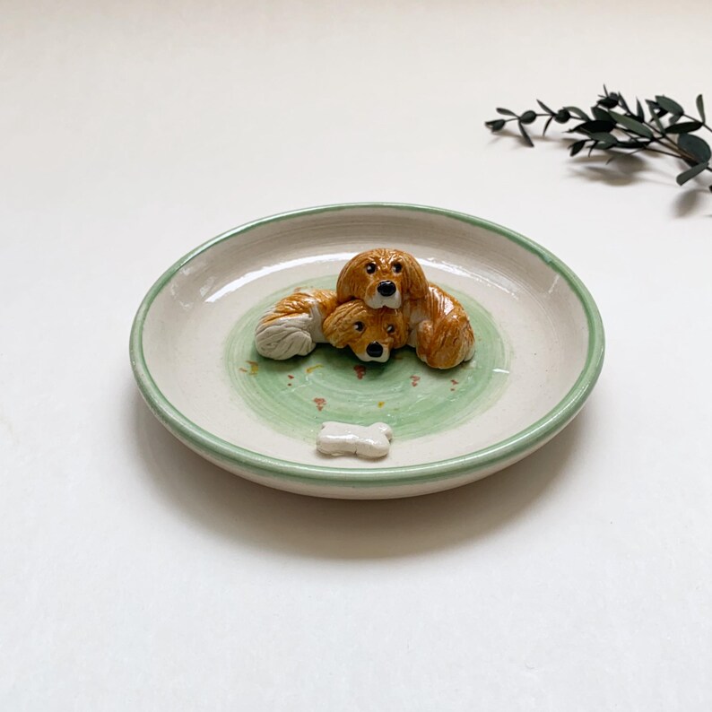 May include: A ceramic dish with a light green rim and a green and white interior. The dish features three brown and white dog figurines and a small bone-shaped accent. A decorative piece for dog lovers.