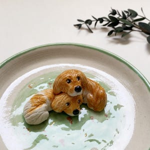 May include: A ceramic plate with a light green and white glaze, featuring two brown and white dog figurines. The dogs are lying on top of each other, with black noses and detailed fur. The plate has a green rim.