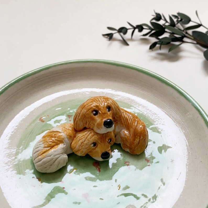 May include: A ceramic plate with a light green and white glaze, featuring two brown and white dog figurines. The dogs are lying on top of each other, with black noses and detailed fur. The plate has a green rim.