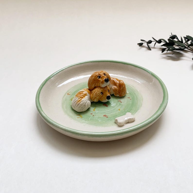 May include: A ceramic dish with a light green rim and a green swirl design in the center. Three small, brown and white dog figurines are arranged on the dish, along with a small bone-shaped figurine.