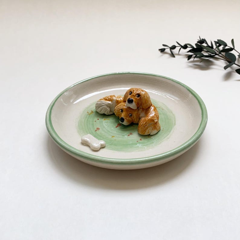 May include: A ceramic dish with a light green rim and a painted green and white interior. The dish features three brown dog figurines and a white bone-shaped accent. The dish is approximately 6 inches in diameter.