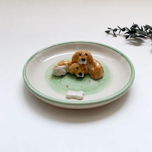 May include: A ceramic plate with a light green rim and a green and white interior. The plate features three brown and white dog figurines and a small white bone. The plate is a decorative item.