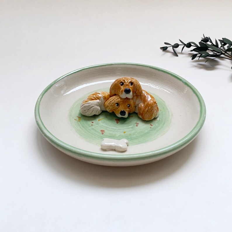 May include: A ceramic plate with a light green rim and a green and white interior. The plate features three brown and white dog figurines and a small white bone. The plate is a decorative item.