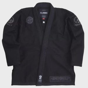 Op de afbeelding: Zwarte vechtsport gi met het woord "CLASSIC" op de kraag. De gi heeft verschillende patches met logo's en de woorden "SHOYOROLL" aan de onderkant. De gi is gemaakt van duurzaam materiaal en is ontworpen voor training en competitie.