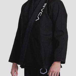 May include: Black martial arts uniform with white logo text on the jacket and pants. The jacket has a wrap-around design with a black belt. The logo text on the jacket reads "RVCA". The pants have a geometric design and the text "VA RVCA".