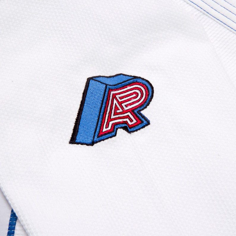 May include: Close-up of white fabric featuring a blue, red, and white embroidered letter "R" patch. The patch has a 3D effect with a blue outline and a red and white interior. The fabric has a textured appearance.
