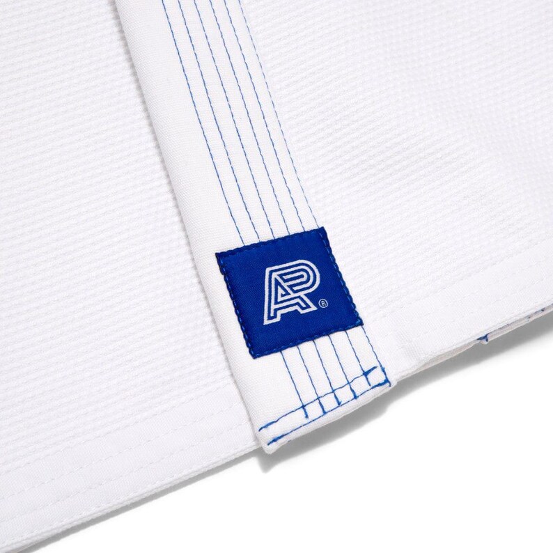 May include: Close-up of a white martial arts uniform with blue stitching and a blue square patch. The patch features a white stylized letter "A" design. The uniform has a textured weave.