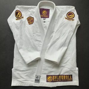 Puede incluir: Un gi de Jiu-Jitsu blanco con parches dorados y morados de la marca "Shoyoroll". El gi tiene cuello y está hecho de tela acanalada. El gi está tendido sobre una superficie oscura.