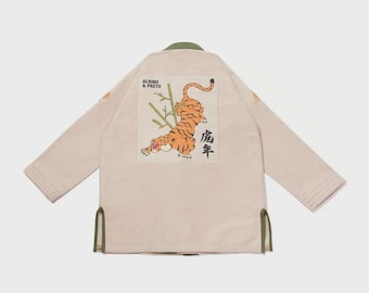 May include: Beige martial arts gi with a tiger illustration on the back. The tiger is orange and black, with bamboo stalks. The gi has green trim and the text "ALBINO & PRETO".