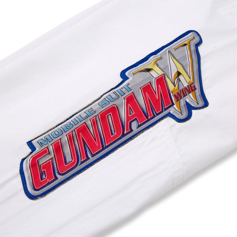 May include: White fabric with a colourful patch featuring the text "MOBILE SUIT GUNDAM WING" in red and silver, outlined in blue. The patch has a star-like design in gold and yellow.