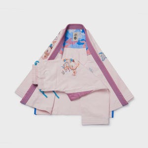 Op de afbeelding: Een lichtroze vechtsportuniformset, bestaande uit een jas en een broek. De jas heeft paarse biezen en een blauw-roze patroon aan de binnenkant. De broek heeft een taille met trekkoord. De set heeft geborduurde bloemen- en drakenontwerpen.