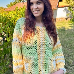 Puede incluir: Un suéter de ganchillo en tonos de amarillo, verde y blanco. El suéter tiene un cuello redondo y mangas largas. El diseño de tejido abierto es visible en toda la prenda. La persona sonríe y lleva el suéter al aire libre.