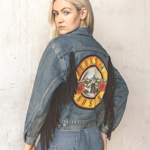 May include: A light-wash denim jacket with a Guns N' Roses logo patch on the back. The jacket features brown fringe detailing on the sleeves and a classic button-up design. The model is wearing blue jeans.