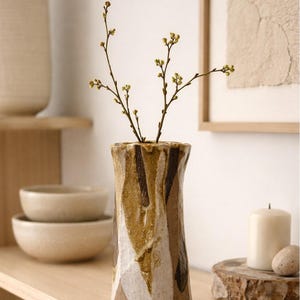 Puede incluir: Un jarrón de cerámica texturizada en tonos marrón, blanco y beige. Contiene delicadas ramas con pequeños capullos verdes. El jarrón se exhibe en una estantería de madera junto con otros elementos decorativos, como cuencos y una vela.
