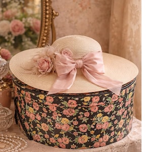 May include: A wide-brimmed straw hat with a large pink bow and floral accents. The hat rests on a round, vintage hat box covered in a black floral pattern. The hat box is approximately 12 inches in diameter.