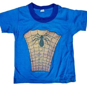 May include: A blue t-shirt with a dark blue collar and sleeves. The shirt features a graphic of a spider and web design in shades of brown and black. The shirt is likely for a child.