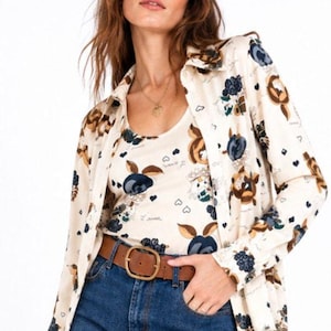 May include: A cream-colored long-sleeve button-down shirt and matching tank top featuring a dark blue and brown floral pattern with small heart icons. The outfit is paired with blue denim jeans and a brown leather belt with a gold-toned buckle.