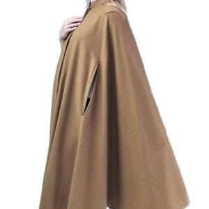 Puede incluir: Una capa de color marrón claro con cuello alto y cierre de botones. La capa presenta un diseño fluido con bolsillos laterales. La modelo lleva pantalones negros y tacones negros. La capa está hecha de una tela suave y fluida.