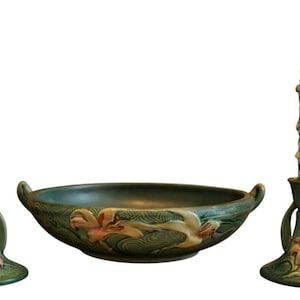 Puede incluir: Un centro de mesa decorativo con un cuenco de cerámica verde con relieve floral y dos candelabros a juego. Los candelabros tienen detalles florales rosas y sostienen velas retorcidas encendidas. El conjunto está diseñado para la decoración del hogar.