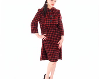 May include: A knee-length dress featuring a red and black plaid pattern, three-quarter length sleeves, and a wide collar with ruffled edges. The bodice includes three large black buttons down the front, creating a structured appearance.