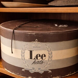 May include: A round, vintage hat box with a brown lid and a striped design in shades of brown, gray, and cream. The box features the text "Lee hats" within a laurel wreath and crown emblem. A black string is tied around the box.