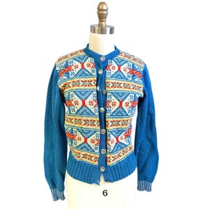 Könnte beinhalten: Blauer Vintage-Strickcardigan mit farbenfrohem geometrischem Muster. Der Pullover hat einen runden Ausschnitt, lange Ärmel und silberne Knöpfe. Das Muster umfasst rote, weiße, gelbe und blaue Designs. Das Kleidungsstück ist auf einer Schaufensterpuppe.