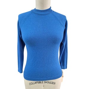 May include: A vibrant blue sweater with a mock neck and long sleeves. The sweater is displayed on a mannequin, showcasing its form-fitting design. The garment features raglan sleeves and a ribbed neckline. The tag reads "COLLAPSIBLE SHOULDER".