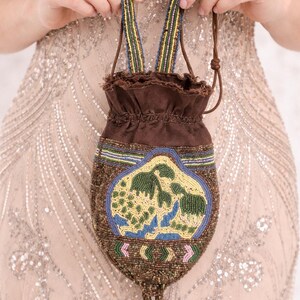 Può includere: Una borsa marrone con coulisse e perline con un design decorativo. La borsa presenta un design centrale con perline verdi, blu e gialle. Le cinghie sono a righe con perline blu, verdi e gialle. La borsa è tenuta da una persona.
