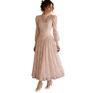 May include: A blush-colored lace dress with long sleeves and a high neckline. The dress features a fitted bodice and a flowing skirt, creating an elegant silhouette. The sleeves have a slight puff at the shoulders and taper to the wrists. The dress is paired with matching heels.