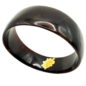 Può includere: Un braccialetto a cerchio marrone scuro con finitura lucida. Il braccialetto ha una fascia larga e una superficie liscia e curva. Un piccolo emblema dorato di una tartaruga è attaccato al braccialetto. Il gioiello è probabilmente in resina.