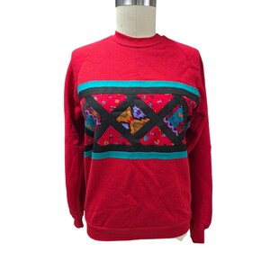 Może przedstawiać: Czerwona bluza z geometrycznym wzorem. Bluza ma okrągły dekolt i długie rękawy. Projekt zawiera czarną, turkusową i wzorzystą tkaninę pośrodku. Geometryczny wzór ma żółte, fioletowe i niebieskie akcenty.