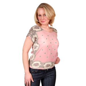 Könnte beinhalten: Ein rosa Oberteil mit kurzen Ärmeln, floralem Muster und dekorativen Verzierungen. Das Oberteil hat einen runden Ausschnitt und wird mit einer blauen Jeans kombiniert. Die Verzierung ist cremefarben mit einem floralen Design.