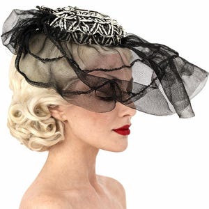 May include: A black and white decorative hat with a veil. The hat features a detailed, patterned design on the crown and a flowing black veil. The hat is a vintage style.