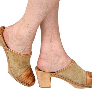 May include: Wooden clogs with tan suede and leather detailing. The clogs feature a block heel and open back design. The toe box is made of a darker brown leather. These neutral-toned clogs could be paired with various outfits.