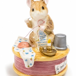 May include: A ceramic figurine of a brown mouse sewing a blue and white striped fabric. The mouse sits on a large pink spool of thread with a silver thimble, a gold candle holder, and a button. A white label on the spool reads No more twist.