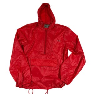 May include: A vibrant red anorak jacket with a hood, half-zip, and a front pocket. It features a drawstring at the hem and a small logo on the chest. The jacket is made from a lightweight material, possibly nylon, and is designed for outdoor wear.