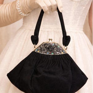 Puede incluir: Un bolso negro vintage con un cierre decorativo con incrustaciones de joyas. El cierre presenta un diseño floral con piedras preciosas de colores. El bolso tiene forma redondeada y asas negras.