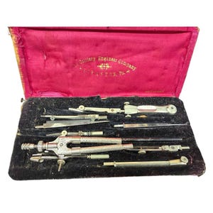 May include: A vintage metal drafting instrument set housed in a rectangular case with a black velvet interior and a red silk-lined lid. The lid features gold text reading The Colliery Engineer Company Scranton, Pa. and Germany.