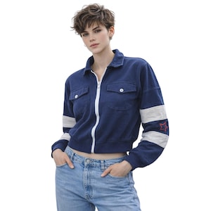 May include: A navy blue cropped jacket with a white zipper and white stripes on the sleeves. The jacket has two front pockets and a red star emblem on the left sleeve. The model is wearing light blue jeans.