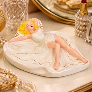 May include: A decorative figurine of a woman in a white dress, reclining on a white base. The figurine has blonde hair and red lipstick. The base is oval-shaped with gold detailing. Other items on the vanity include perfume bottles and a mirror.