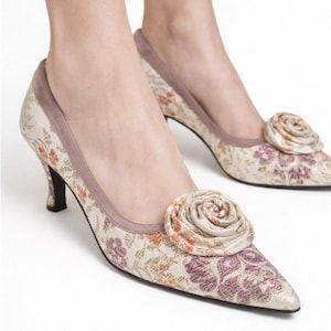 Op de afbeelding: Pumps met bloemenprint, een spitse neus en een kleine hak. De schoenen hebben een grote stoffen roos op de neus en een gedempt kleurenpalet van crème, paars en oranje. De hakken zijn ongeveer 5 cm hoog.