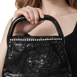 Puede incluir: Bolso de mano negro con lentejuelas con la parte superior redondeada y un asa negra. El bolso está adornado con pequeñas lentejuelas redondas y una fila de pequeños diamantes de imitación blancos a lo largo del borde superior. El bolso lo sostiene una persona.