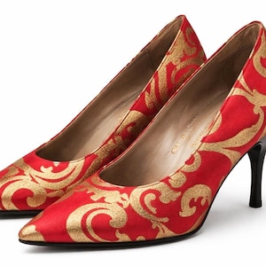 Puede incluir: Un par de zapatos de tacón rojos con un estampado floral dorado. Los zapatos tienen punta y tacones negros. Los tacones miden aproximadamente 7,6 cm de alto.