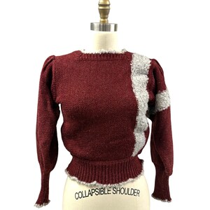 May include: A maroon sweater with a white and gray knit design on the right shoulder. The sweater has a ribbed hem and cuffs. "COLLAPSIBLE SHOULDER" is written on the mannequin's torso.