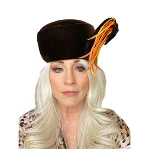 Può includere: Un cappello a pillola in velluto marrone con un accento di piuma gialla e arancione.