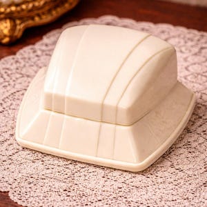 Puede incluir: Caja de baratijas de color marfil, estilo Art Déco, con un diseño geométrico escalonado. La caja está sobre un delicado tapete de encaje color crema, lo que sugiere una estética vintage. La tapa tiene ranuras verticales.