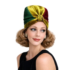 Puede incluir: Un turbante de terciopelo en verde, dorado y burdeos. El turbante está anudado en la parte delantera y tiene un aspecto suave y lujoso. El sombrero lo lleva una persona que lleva un vestido negro y pendientes dorados.