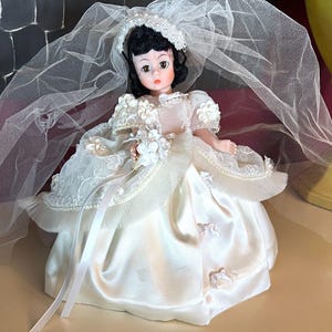 May include: A vintage bride doll wearing a white satin wedding dress with lace and floral accents. The doll has black hair, a veil, and a pearl-adorned headpiece. The doll is holding a bouquet of white flowers.