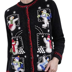 May include: Black cardigan sweater with red trim and button closure. The sweater features embroidered snowmen in various poses, with details like scarves and hats. White snowflakes are scattered around the snowmen. A festive winter-themed garment.